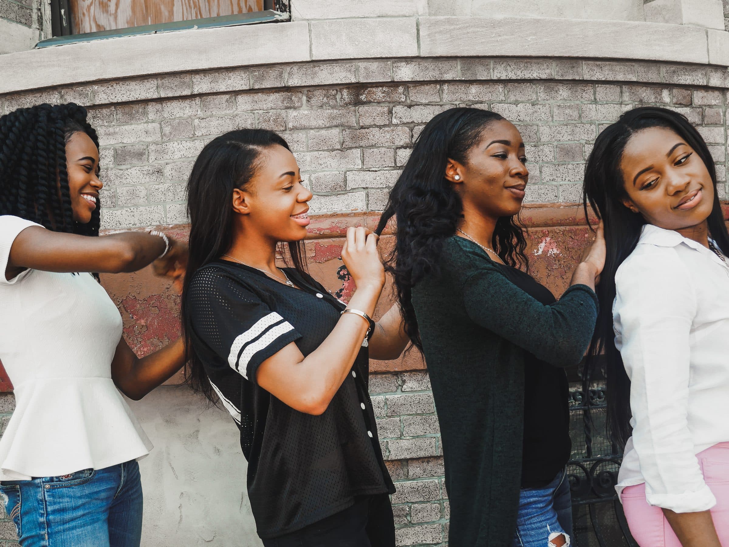 Four young women stand in a line along a brick wall, smiling as each braids the hair of the woman in front of her.