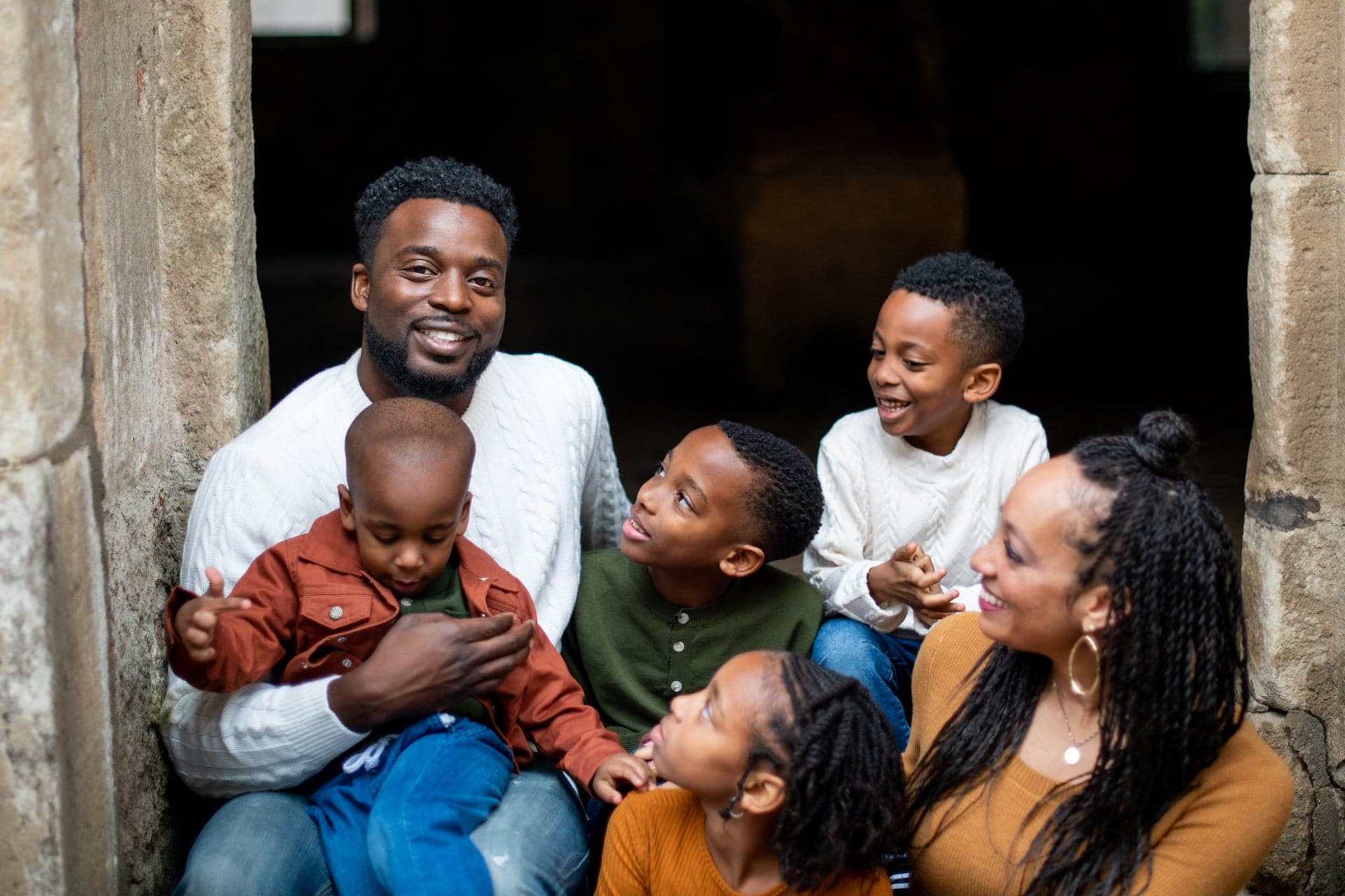 A man and a woman sit close together with four children in a stone doorway, laughing.